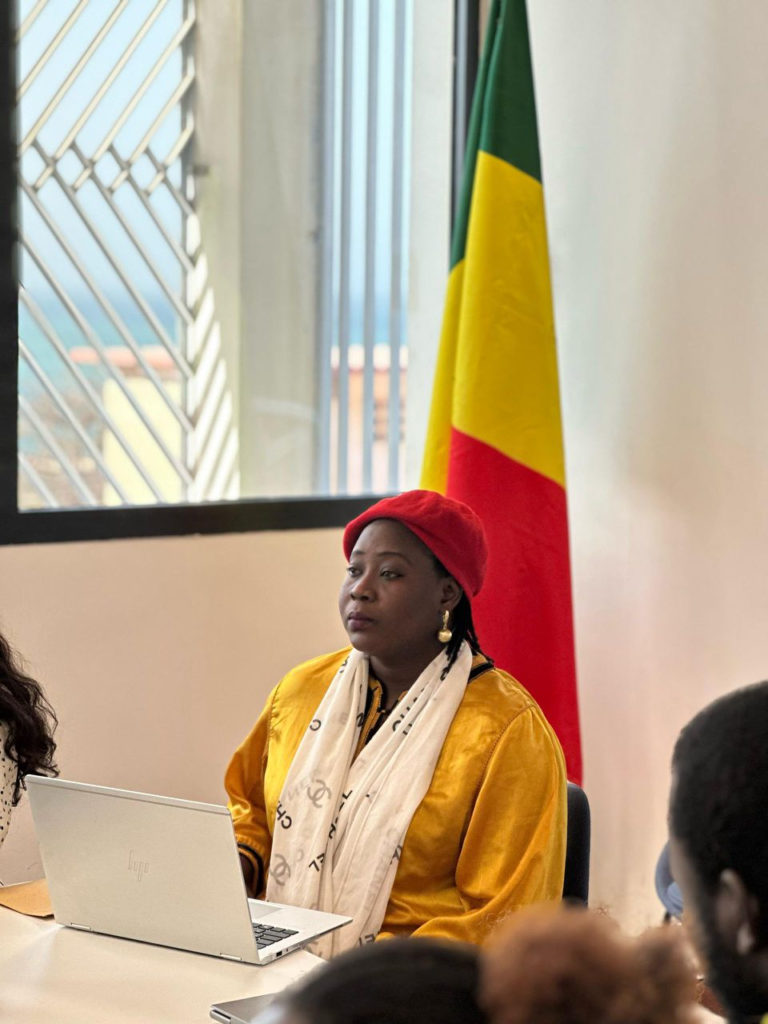Fatou Kiné Ndiaye sitting at a conference table with an open laptop, wearing a yellow top and a red beret, with the Senegal flag in the background.