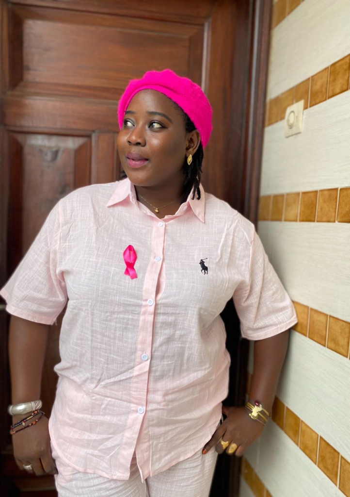 A close-up portrait of Fatou Kiné Ndiaye looking directly at the camera with a bold, determined expression, wearing a pink shirt and a pink beret